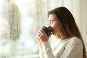 Side view portrait of a relaxed teen drinking coffee looking outside through a window in a rainy day of winter at home